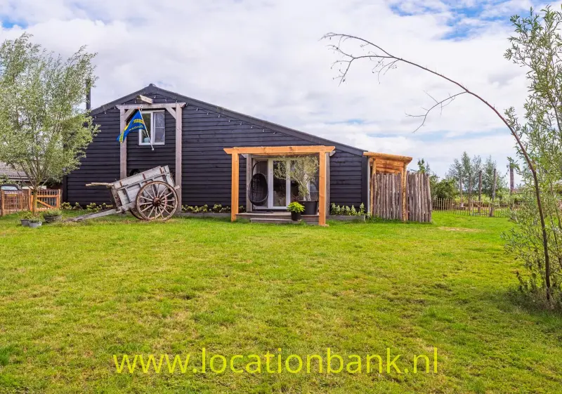 Donker houten gebouw met overdekte veranda, houten kar en ruime groene tuin onder een bewolkte hemel.