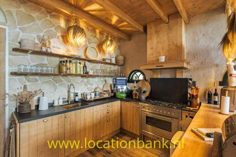 Keuken met stenen wand, houten plafond, kookfornuis, open planken en gevlochten hanglampen.