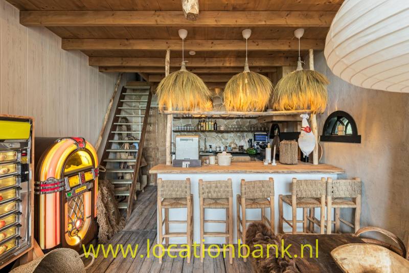 Houten bar met barkrukken, drie gevlochten hanglampen en een kleurrijke jukebox onder houten balken.