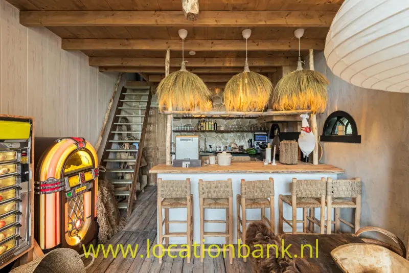 Houten bar met barkrukken, drie gevlochten hanglampen en een kleurrijke jukebox onder houten balken.