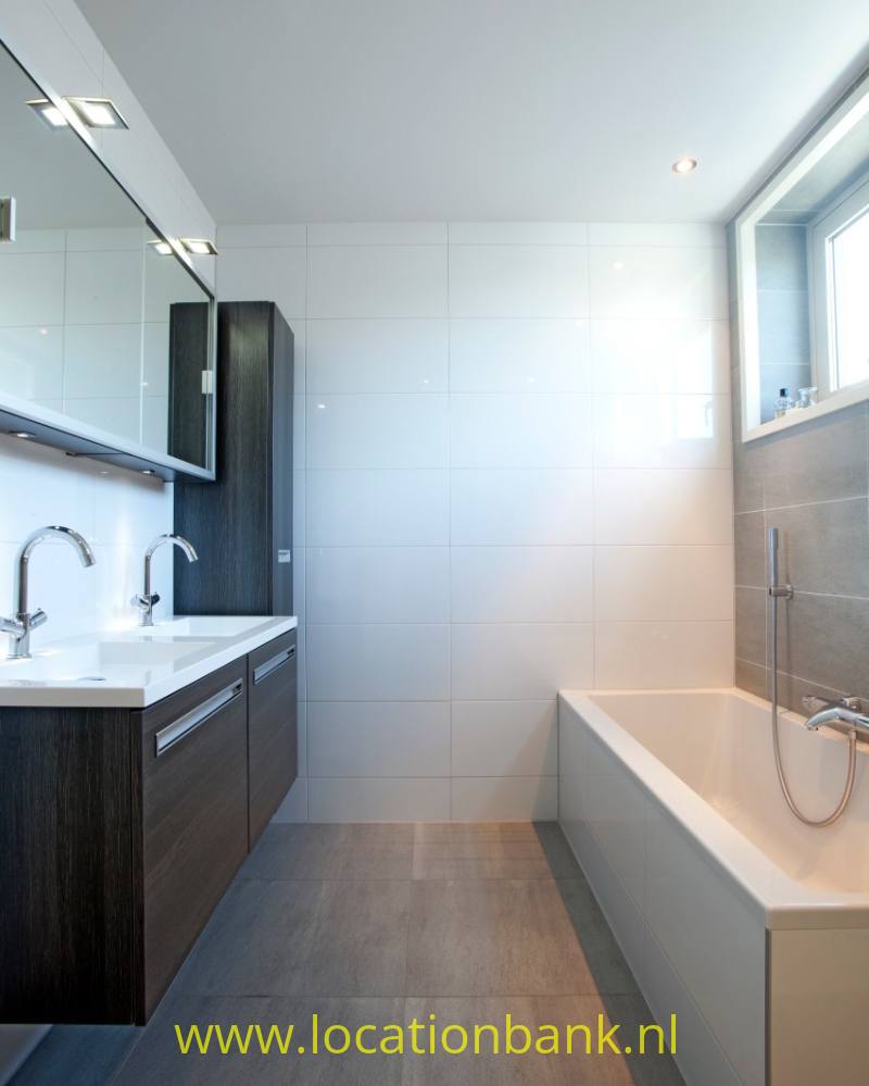 Badkamer met dubbele wastafel, donkere onderkasten, witte wandtegels en een ligbad bij het raam.