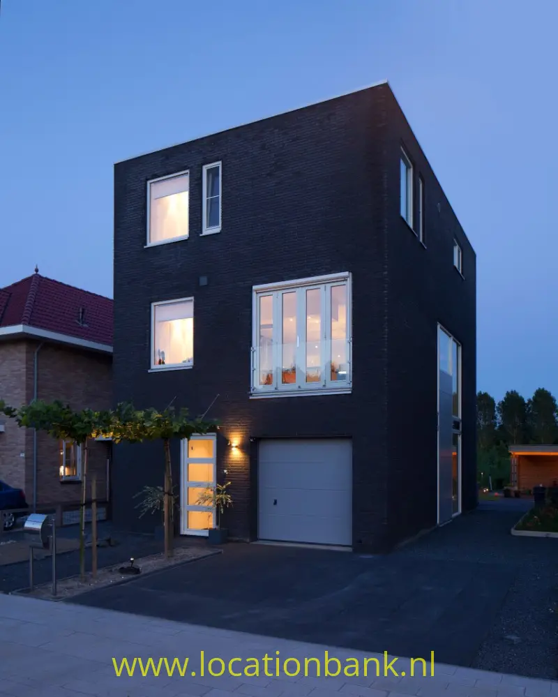 Donker bakstenen vinex hoekhuis met grote ramen, een garage en een verlichte entree aan de straat.