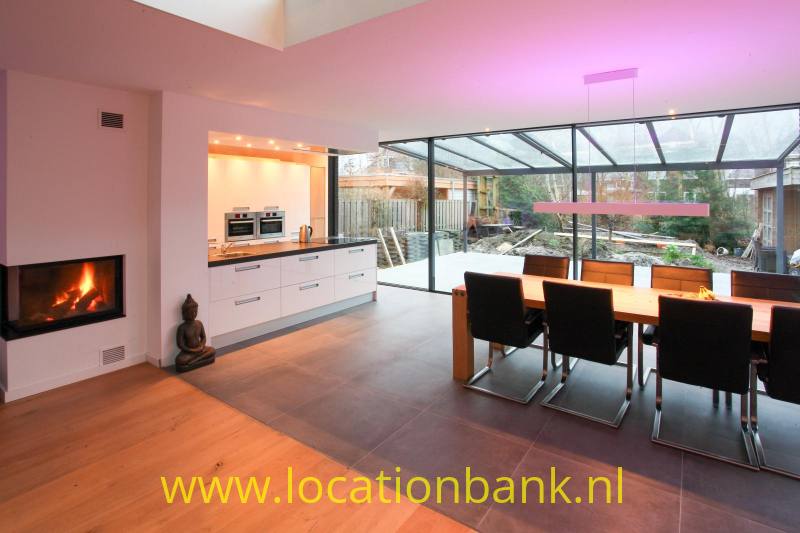 Open woonruimte met witte keuken, eettafel, glazen aanbouw en ingebouwde haard.