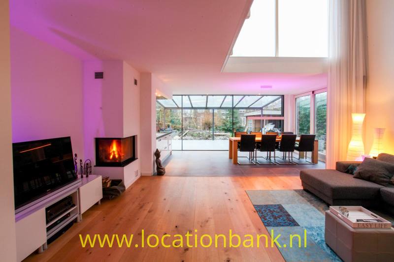 Woonkamer met houten vloer, twee haarden, een hoekbank en een glazen uitbouw.