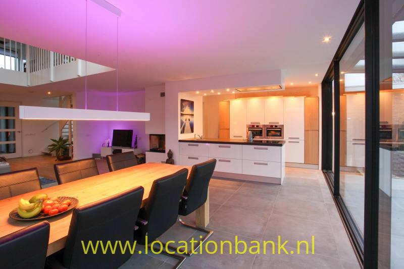 Grote houten eettafel met zwarte stoelen staat voor een witte keuken met kookeiland en glazen deuren.