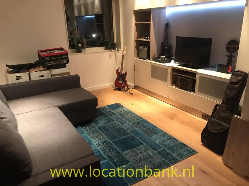 Woonkamer met een grijze hoekbank, blauw kleed, televisie, gitaar en vitrinekast.