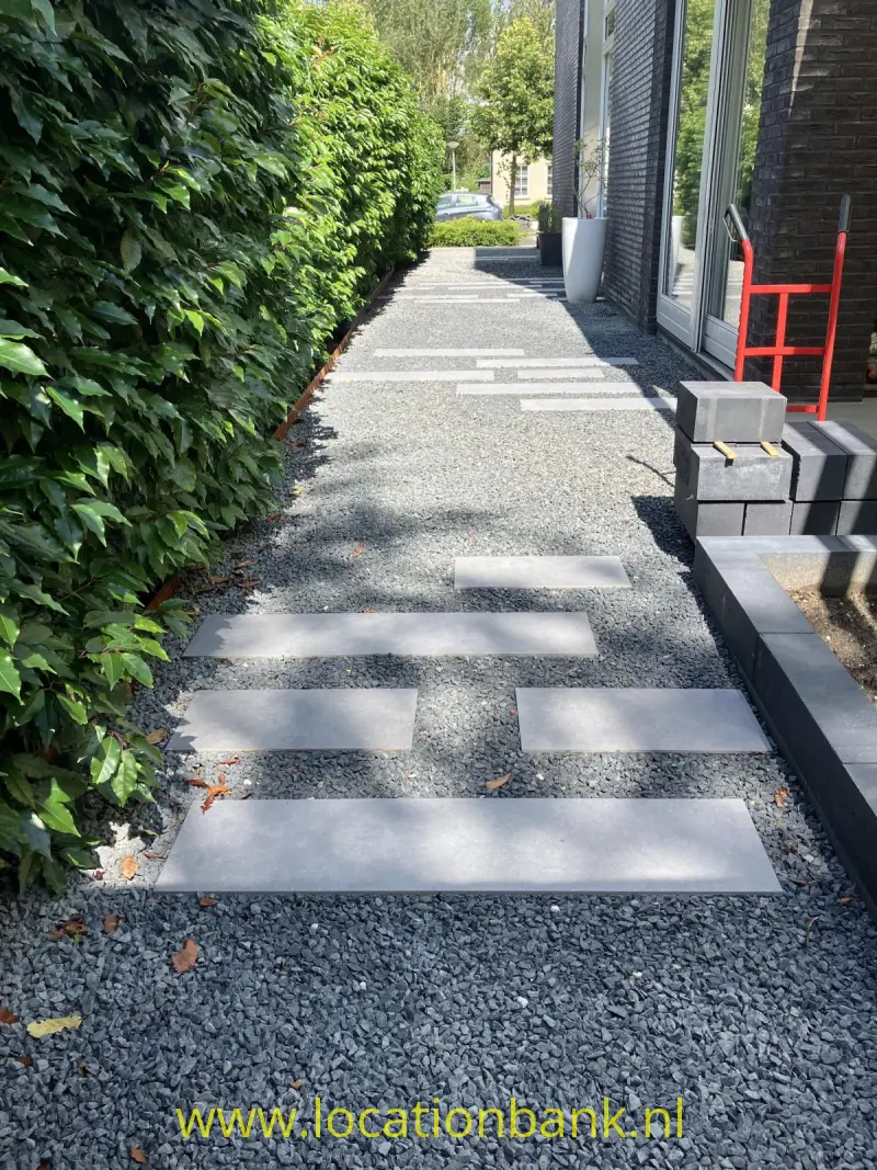 Een grindpad met rechthoekige stapstenen loopt langs een hoge haag en een gebouw.