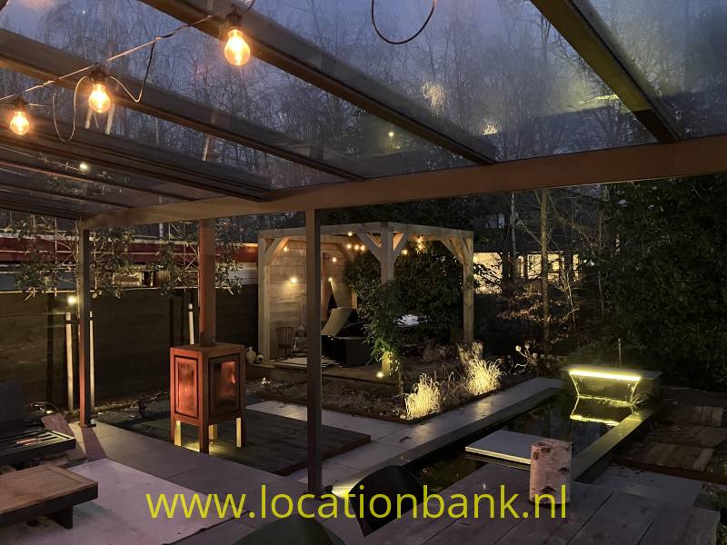 Overdekt terras met lichtslingers, een vijver en verlichte beplanting in een tuin.