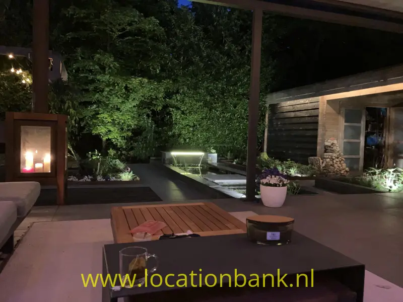 Overdekt terras met een lage tafel, glazen en een verlichte tuin met een langwerpige vijver.