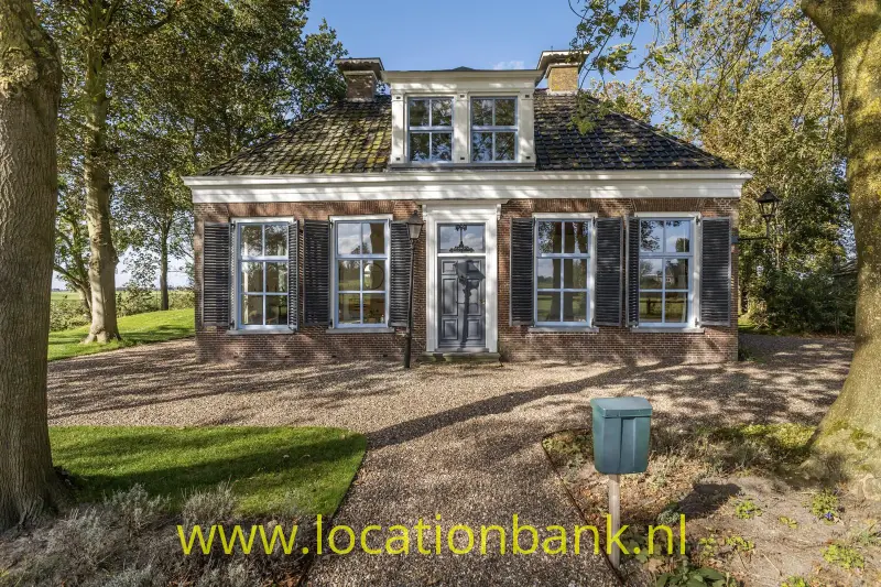 Vrijstaand bakstenen woonboerderij met donkere luiken, een centrale voordeur en een grindpad tussen grote bomen.