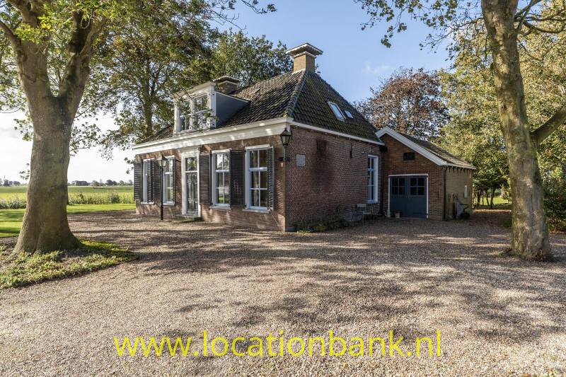 Bakstenen huis met een donker pannendak, witte luiken, een aangebouwde garage en grindoprit tussen bomen en velden.