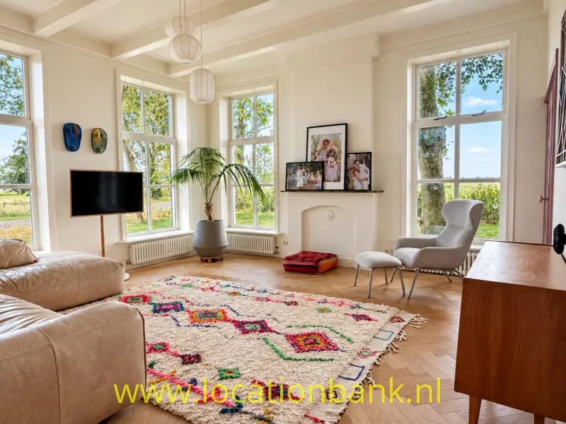 Lichte woonkamer met grote ramen, een kleurrijk vloerkleed met patroon, beige zitmeubels en een houten dressoir.
