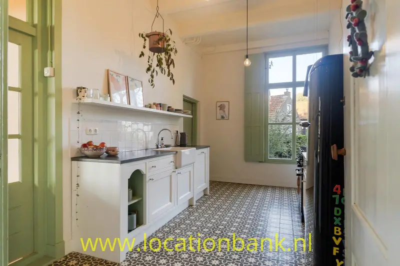 Witte keuken met groene deur en raamkozijn, hangplanten en een tegelvloer met een geometrisch patroon.