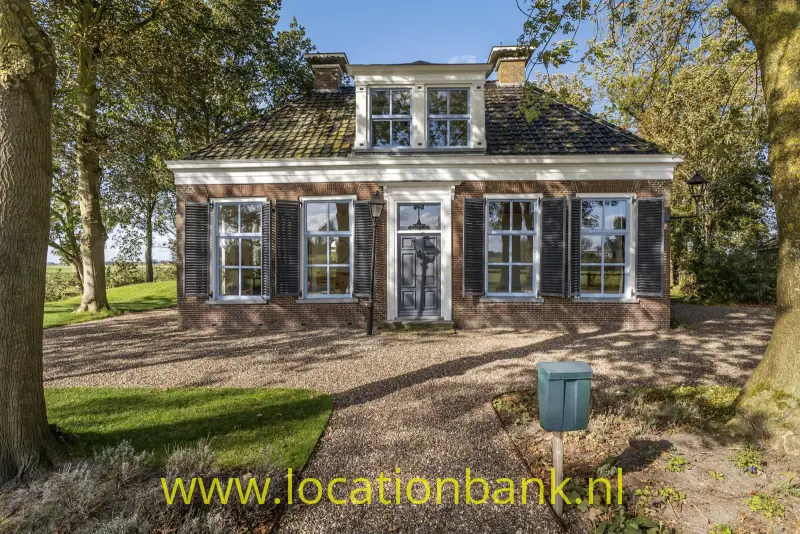 Bakstenen woning met donkere luiken, een centrale voordeur, dakkapel en grindpad tussen de bomen.