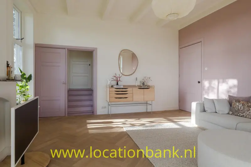 Woonkamer met roze deuren, een houten visgraatvloer, een ronde spiegel en een witte bank.