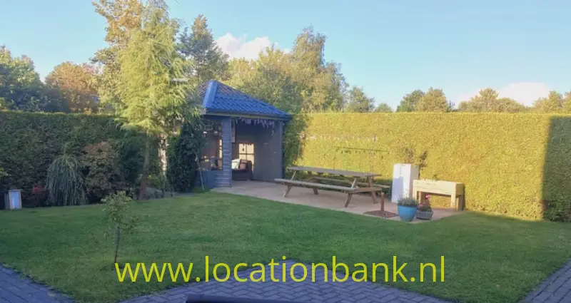 Houten tuinhuisje met overkapping, picknicktafel en bloempot in een tuin met een hoge haag.