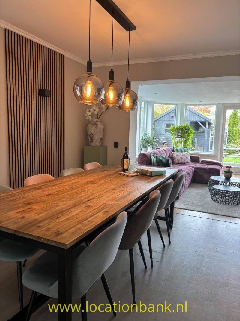 Houten eettafel met grijze stoelen en drie hanglampen staat naast een lichte zithoek met grote ramen.