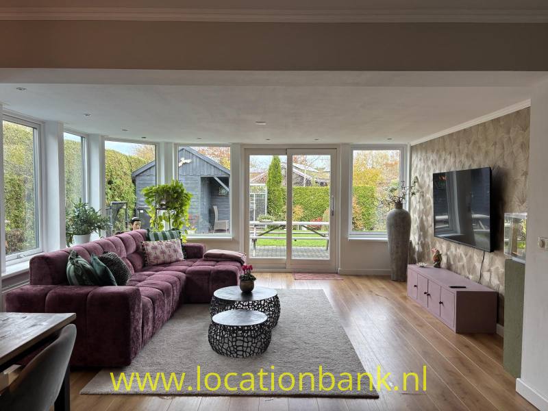 Woonkamer met een paarse hoekbank, grote ramen, een vloerkleed en een televisie aan de wand.