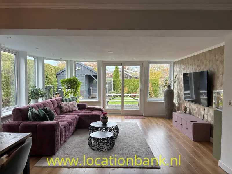 Woonkamer met een paarse hoekbank, grote ramen, een vloerkleed en een televisie aan de wand.