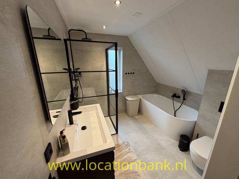 Badkamer met een vrijstaand wit bad, een wastafelmeubel en een zwart omlijnde glazen douchewand.