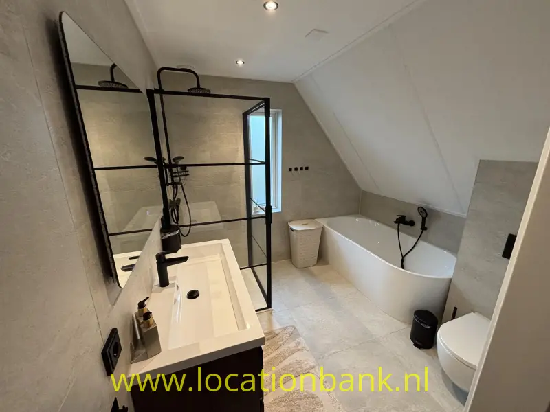 Badkamer met een vrijstaand wit bad, een wastafelmeubel en een zwart omlijnde glazen douchewand.