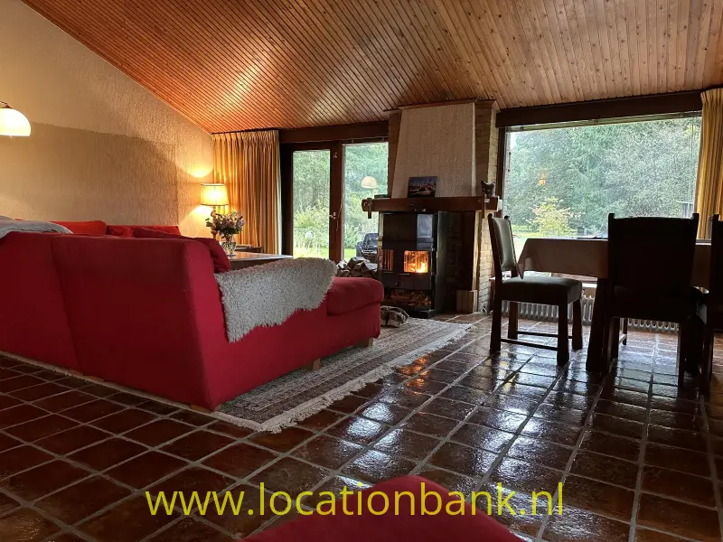 Woonkamer met rode banken, houten plafond, open haard, eettafel en grote ramen met uitzicht op bomen.