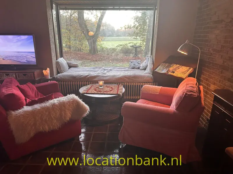 Woonkamer met twee rode fauteuils, een grote raampartij, een zitbank en uitzicht op een groene tuin.