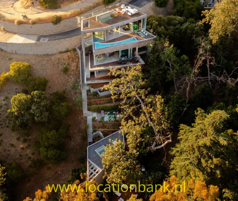 Modern huis met meerdere terrassen en een turquoise zwembad tussen bomen op een steile, droge helling.