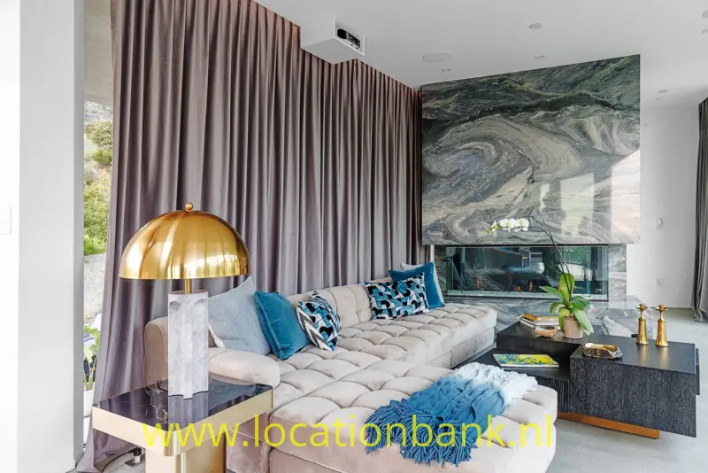 Lichtgekleurde hoekbank met blauwe kussens en plaid staat voor een grote abstracte wanddecoratie in een woonkamer.