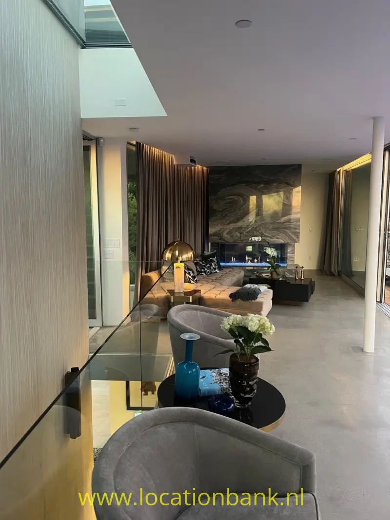 Een grijze fauteuil staat naast een glazen balustrade in een open interieur met zitmeubels en een wandkunstwerk.