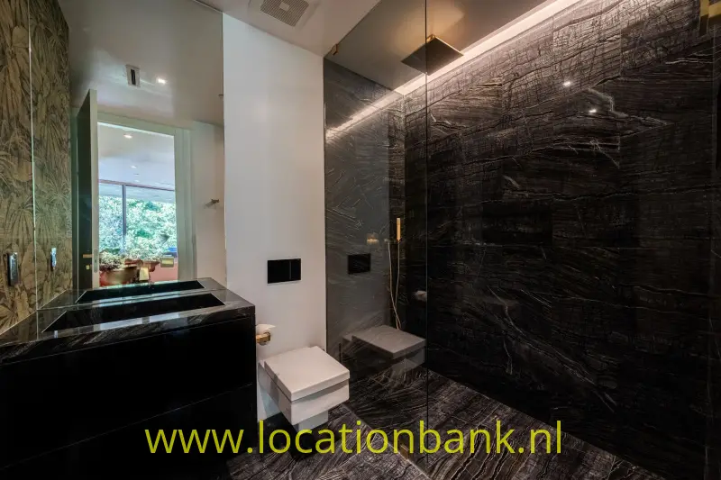 Donkere badkamer met een zwart wastafelmeubel, hangtoilet, glazen douchewand en sterk geaderde wand- en vloerbekleding.
