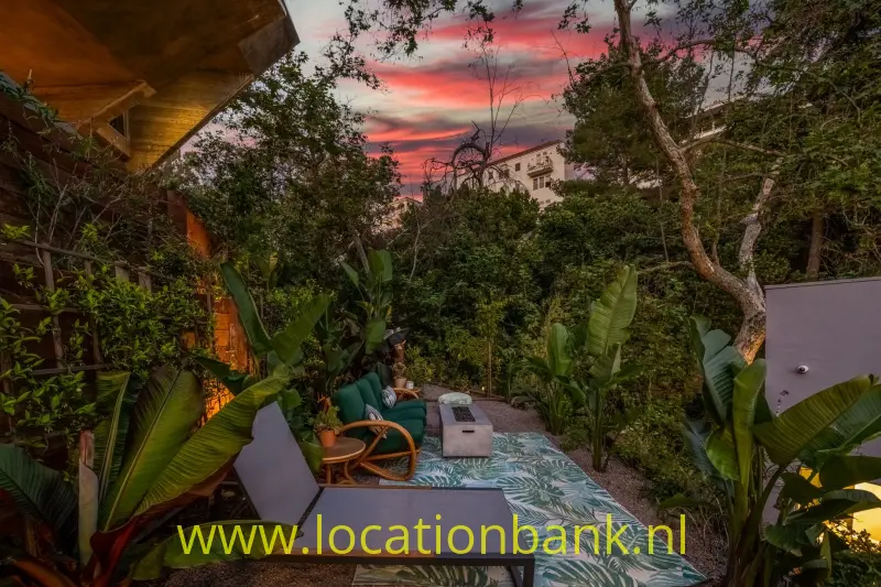 Tropische tuin met ligstoelen, een tegelpad met bladmotief en een rood gekleurde avondlucht.