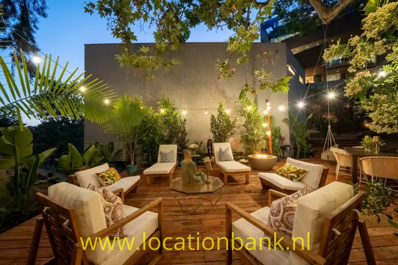Houten terras met banken, fauteuils, potplanten en een centrale vuurkorf tussen de bomen.