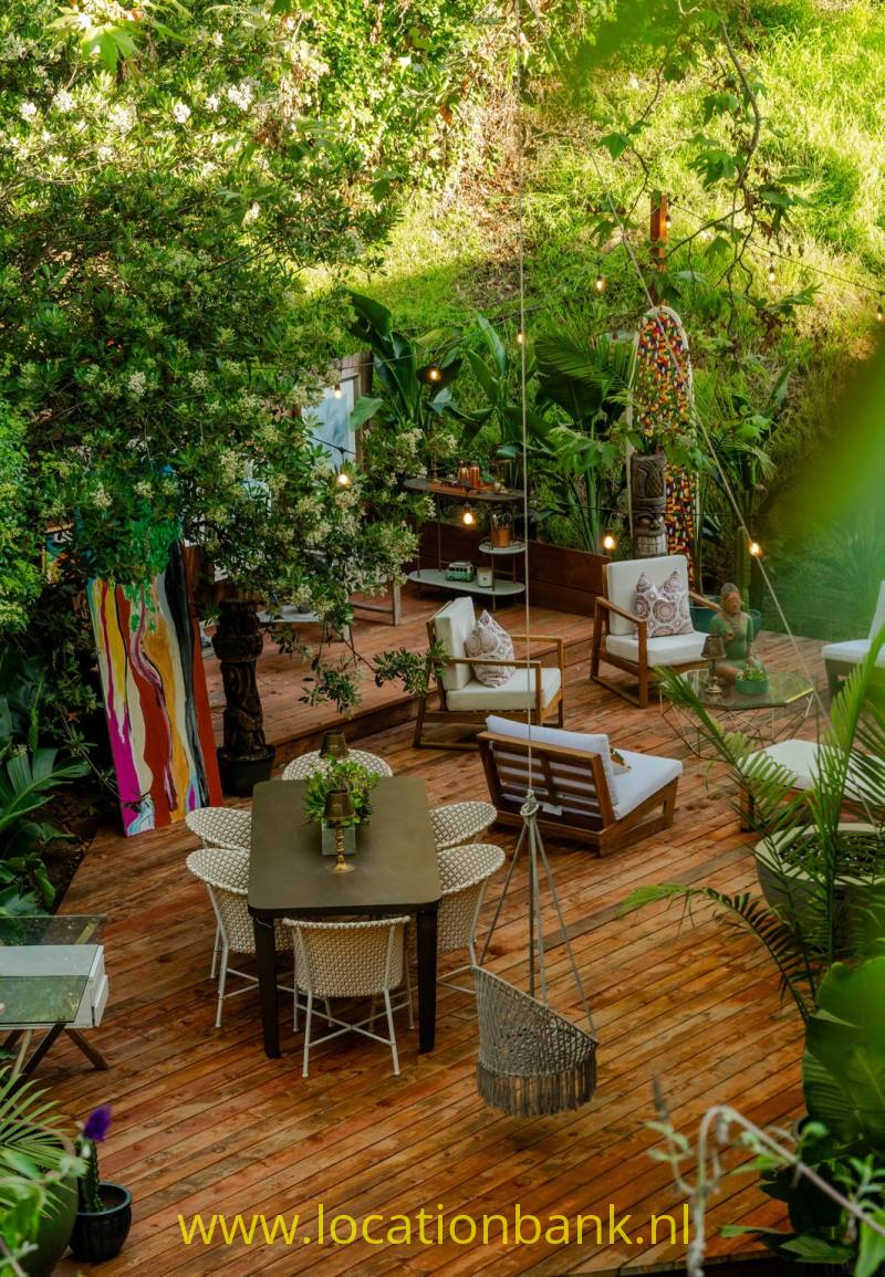 Een houten terras tussen dichte beplanting heeft een eettafel, stoelen en twee loungestoelen.