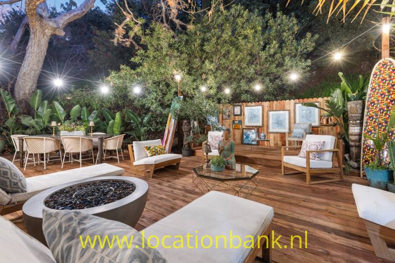 Een houten terras met witte loungestoelen, een ronde schaal met donkere stenen en lichtslingers tussen de bomen.