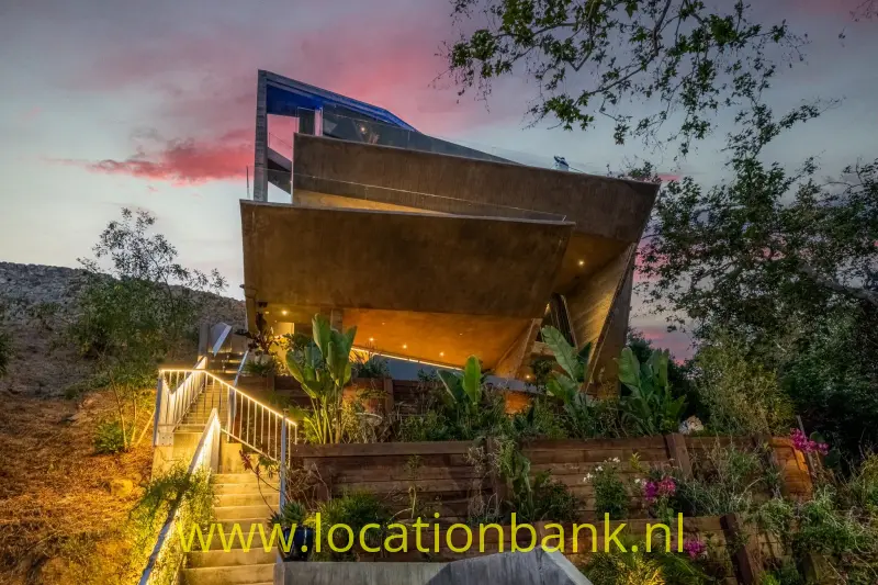 Hoekige villa met overkragende terrassen, een verlichte buitentrap en dichte beplanting tegen een roze avondlucht.