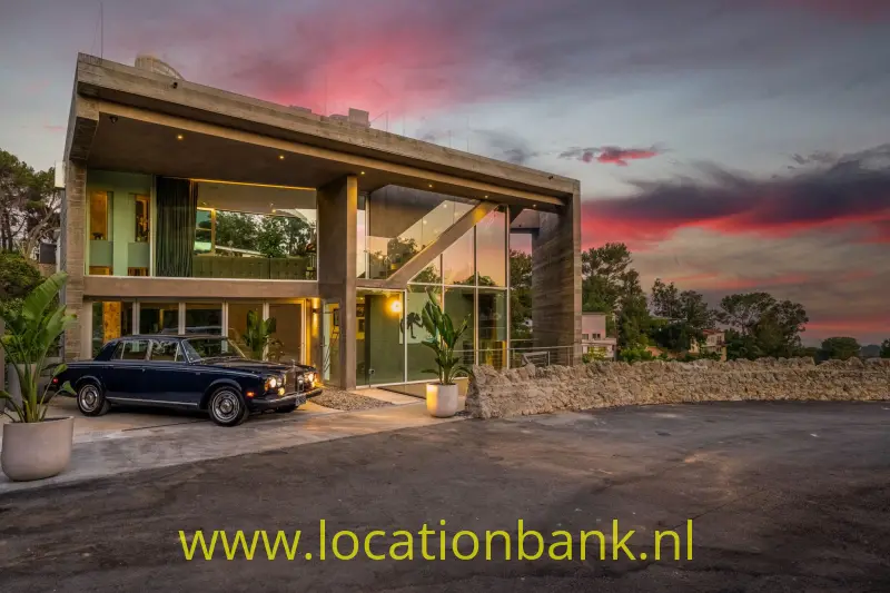 Moderne villa in Los Angeles  met twee verdiepingen, een glazen gevel en een donkere klassieke auto bij zonsondergang.