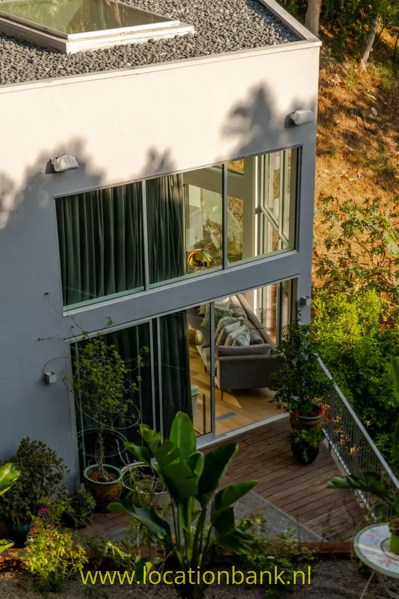 Witte gevel met grote ramen, groene gordijnen, een houten terras en beplanting langs de woning.