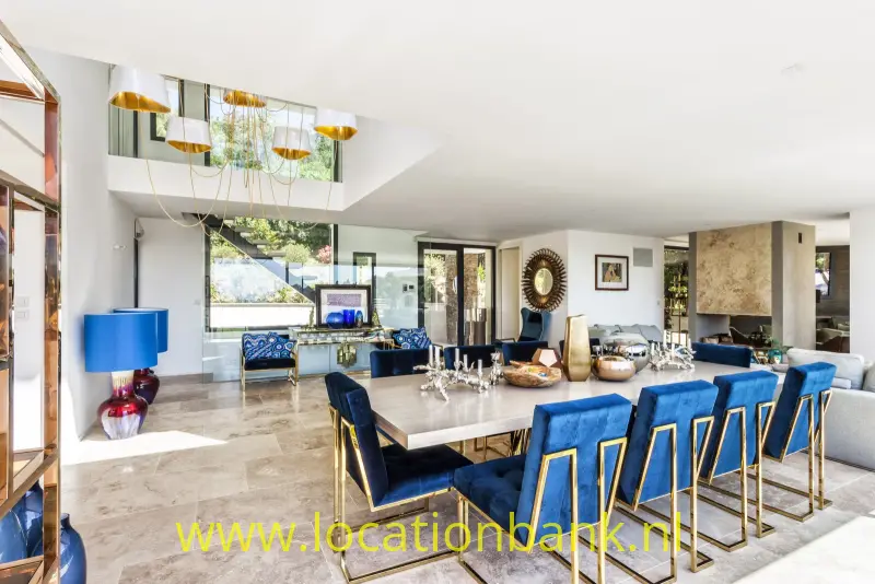 Eetkamer met een lange tafel, blauwe stoelen, glazen deuren en meerdere hanglampen.