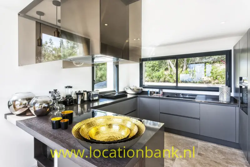 Keuken met een kookeiland, donkere kasten, een grote goudkleurige schaal en brede ramen.