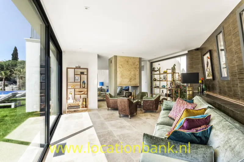 Woonkamer met een groenige bank, twee fauteuils, open wandrekken en hoge glazen schuifdeuren.