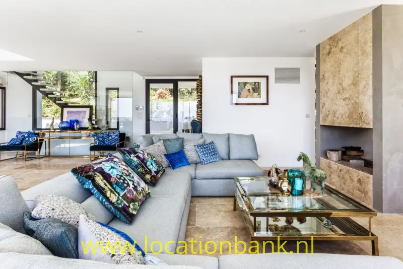 Woonkamer met een grijze hoekbank, glazen salontafel, kleurrijke kussens en een open haard.