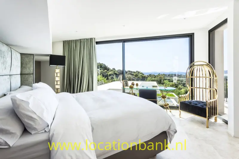 Slaapkamer met een opgemaakt bed, groene gordijnen, grote ramen en een hangstoel met uitzicht.