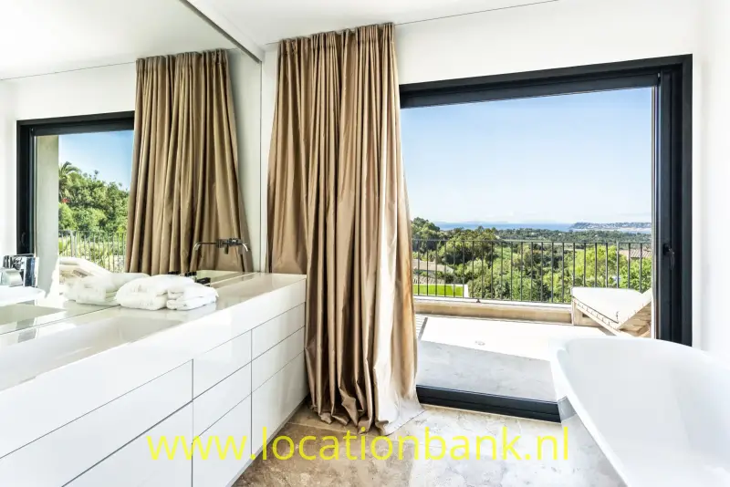 Badkamer met vrijstaand wit bad, dubbele wastafel, beige gordijnen en uitzicht over een groen landschap.