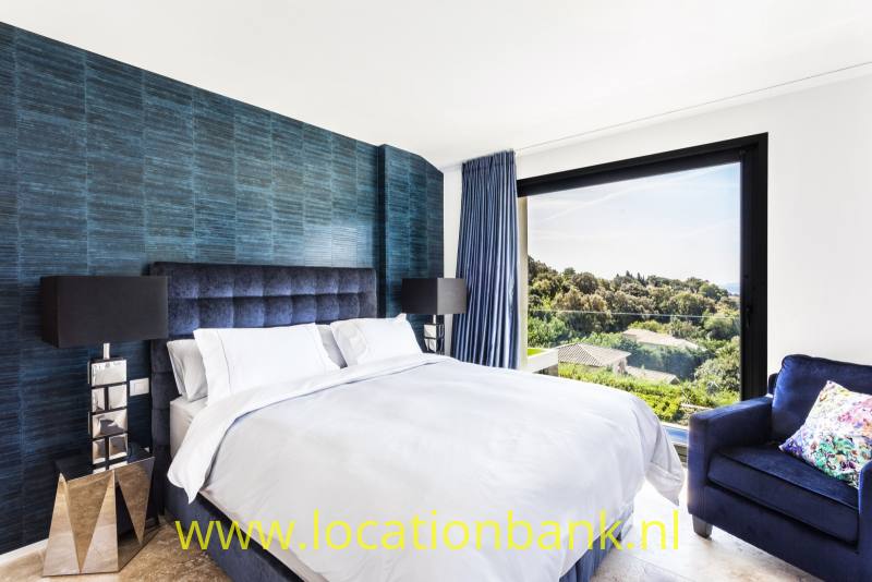 Slaapkamer met een groot wit bed, een donkerblauwe wand met reliëf en een breed raam met uitzicht op groen.