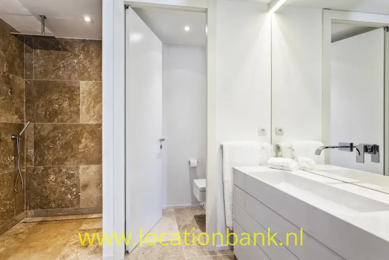 Badkamer met een bruine betegelde inloopdouche, een wit wastafelmeubel en een grote spiegel.
