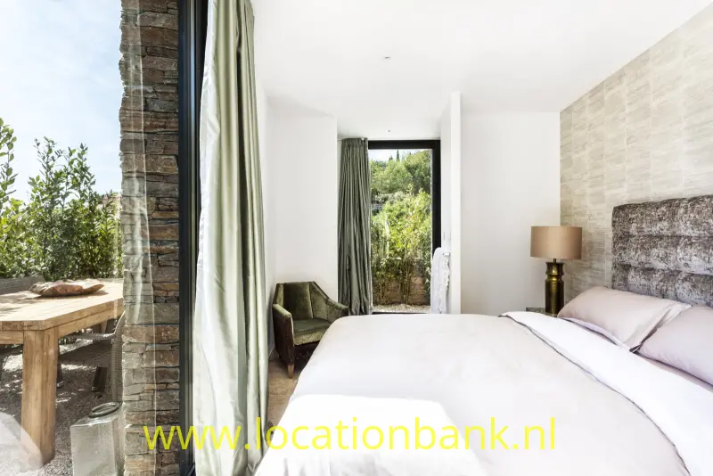 Slaapkamer met een wit bed, een natuurstenen wand, een glazen deur naar buiten en een houten tafel.