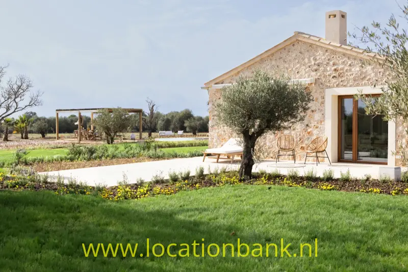 Stenen huis met een olijfboom, bloemperk en gazon, met velden en een houten pergola op de achtergrond.