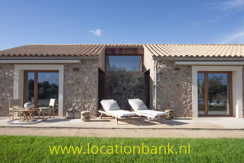 Stenen spaanse villa met terracotta dakpannen, glazen deuren, terrasmeubilair en twee ligstoelen aan een groen gazon.