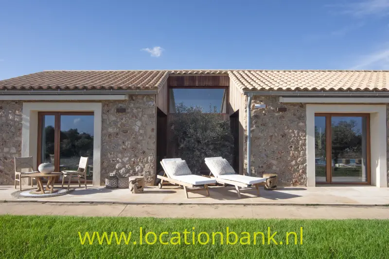 Stenen spaanse villa met terracotta dakpannen, glazen deuren, terrasmeubilair en twee ligstoelen aan een groen gazon.